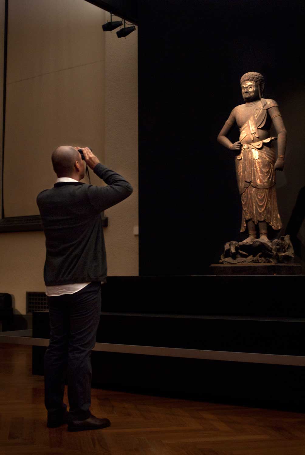 不動明王立像（平安時代・12世紀、東京国立博物館蔵）。 『こういった細かい紋様や、遠くに置かれた仏像を鑑賞する際は、双眼鏡が便利です。 これは不動明王という仏像ですが、先の帝釈天に比べると、厚みも少なく、穏やかな雰囲気で作られているなど、様式の変化がよく分かります。この様式の変化は、社会情勢や信仰のあり方の変化によって、その当時の人々の好みが変わっていったことに由来します。 また、分業化も進んでいたので、この像も、何人かの手で作られていったと思われます。 この像の大きな特徴は「目」。玉眼という、水晶の目が入れられています。本来、この時代には、この玉眼を入れることはなく、その後の時代に修復を受けた際、仏師が改造してしまったのだと想像できます。顔の一部が白っぽくなっているのですが、おそらくここから割られ、内側をくりぬいて玉眼が入れられたのでしょう』。